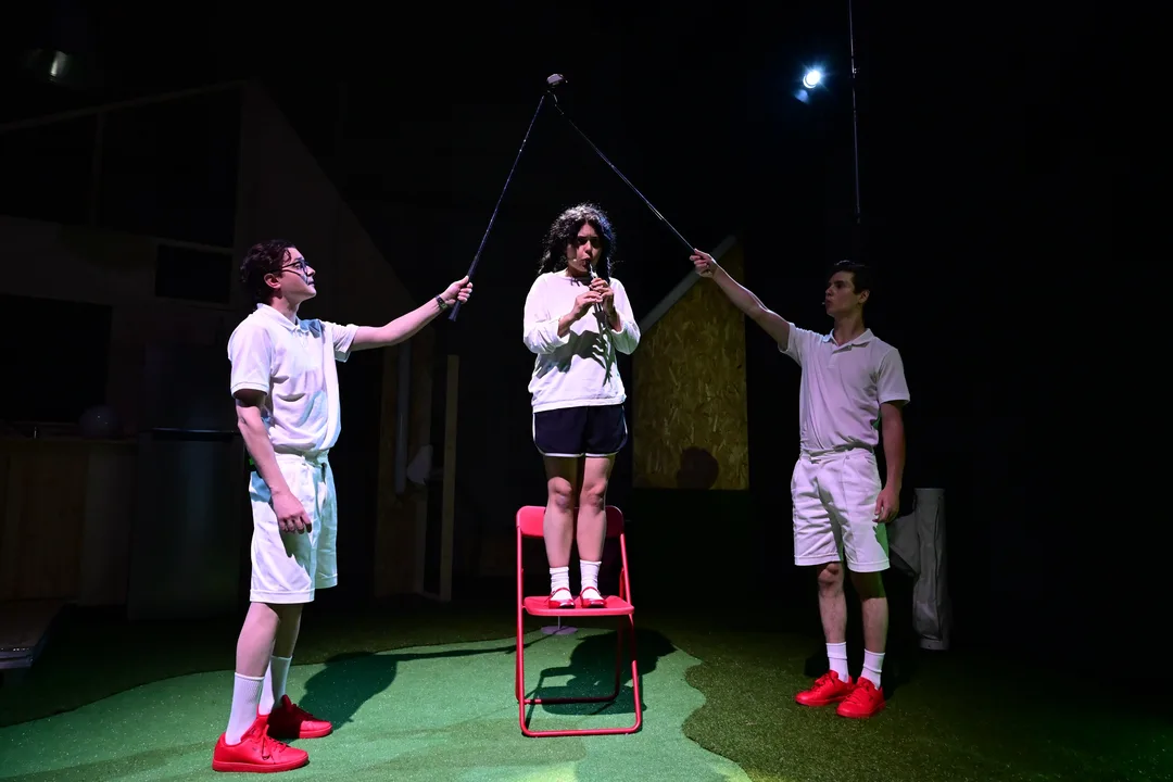 Funny Games: doi băieți în alb țin o crosă deasupra unei fete pe un scaun roșu, pe gazon