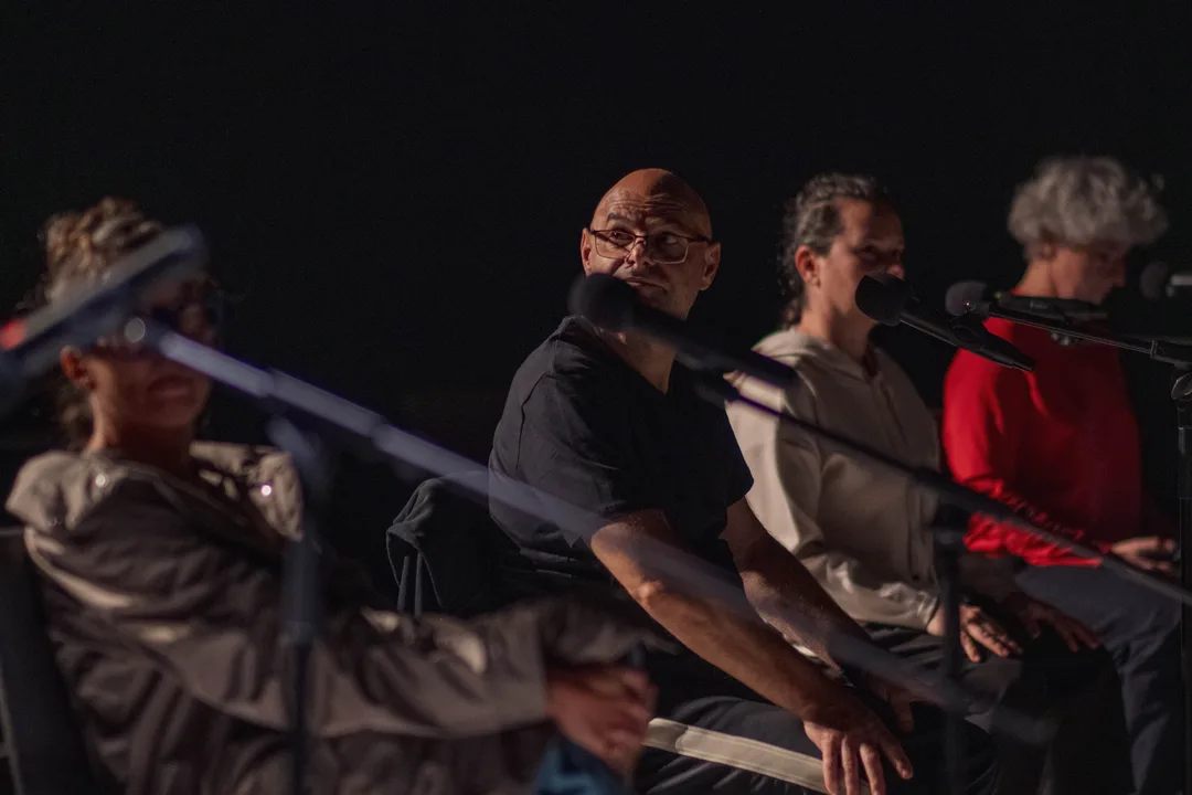 Performerii la microfoane, pe scaune, în semiîntuneric