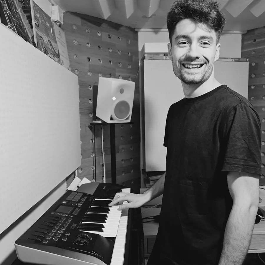 Un om zâmbește la un pian electric, în fața unui panou acustic; alb-negru